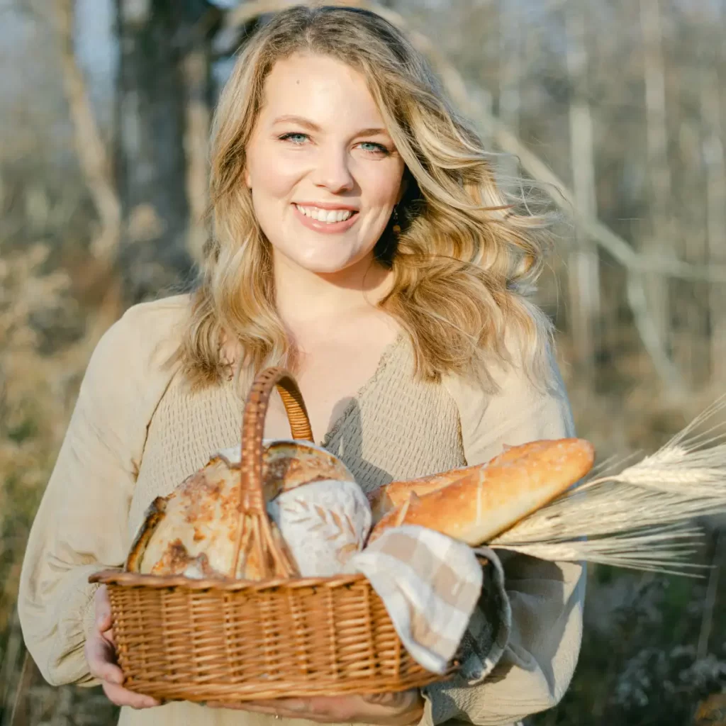 girl in a beige dress standing in a field with trees behind with a basket of bread