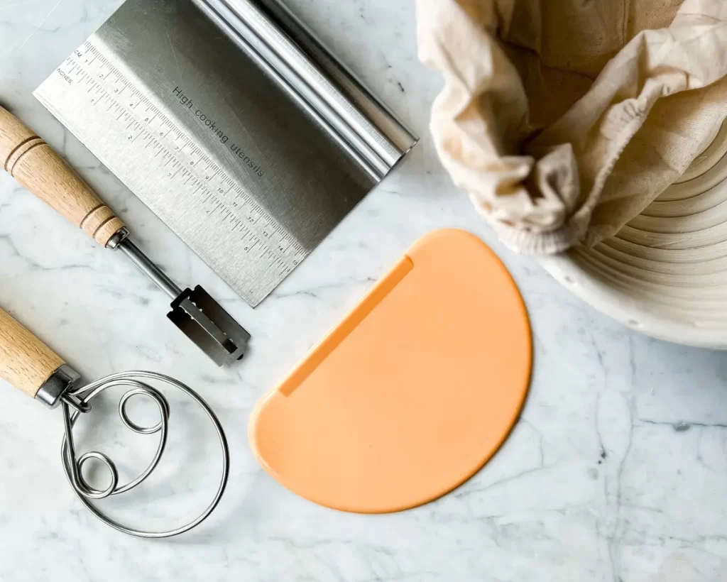 overlay shot of sourdough tools: dough whisk, bread lame, bench and bowl scraper and banneton basket
