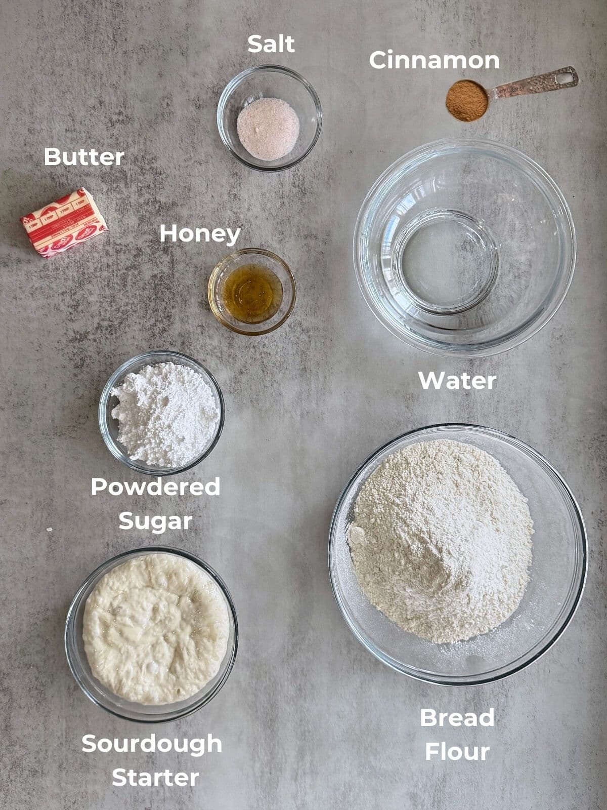Ingredients for cinnamon honey butter sourdough bread on a gray countertop with all of the ingredients in glass bowls.