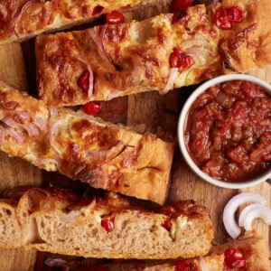 Sourdough salsa focaccia sliced in strips and placed on a wood cutting board with red onions scattered and a bowl of salsa.