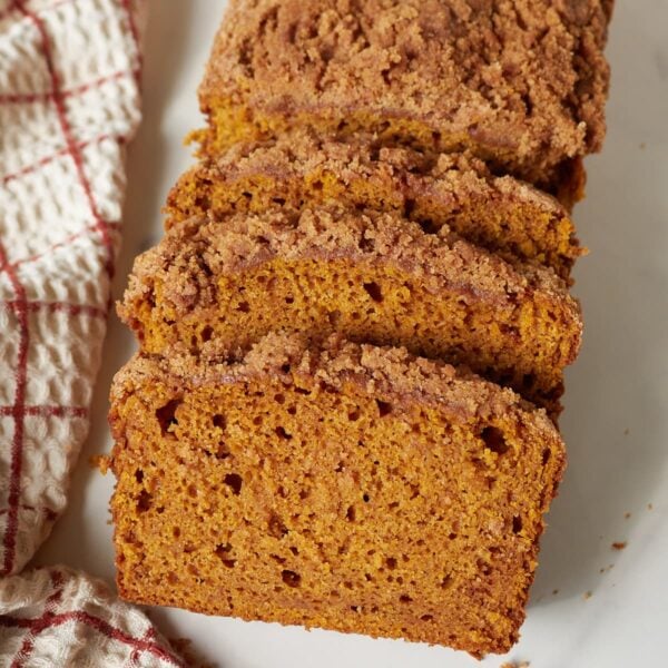 Slices of sourdough pumpkin quick bread on a white board next to a checkered towel.
