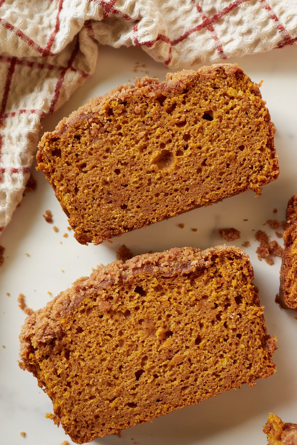 Two slices of sourdough pumpkin quick bread with a streusel topping on a white counter with a cream and red checkered towel next to it.