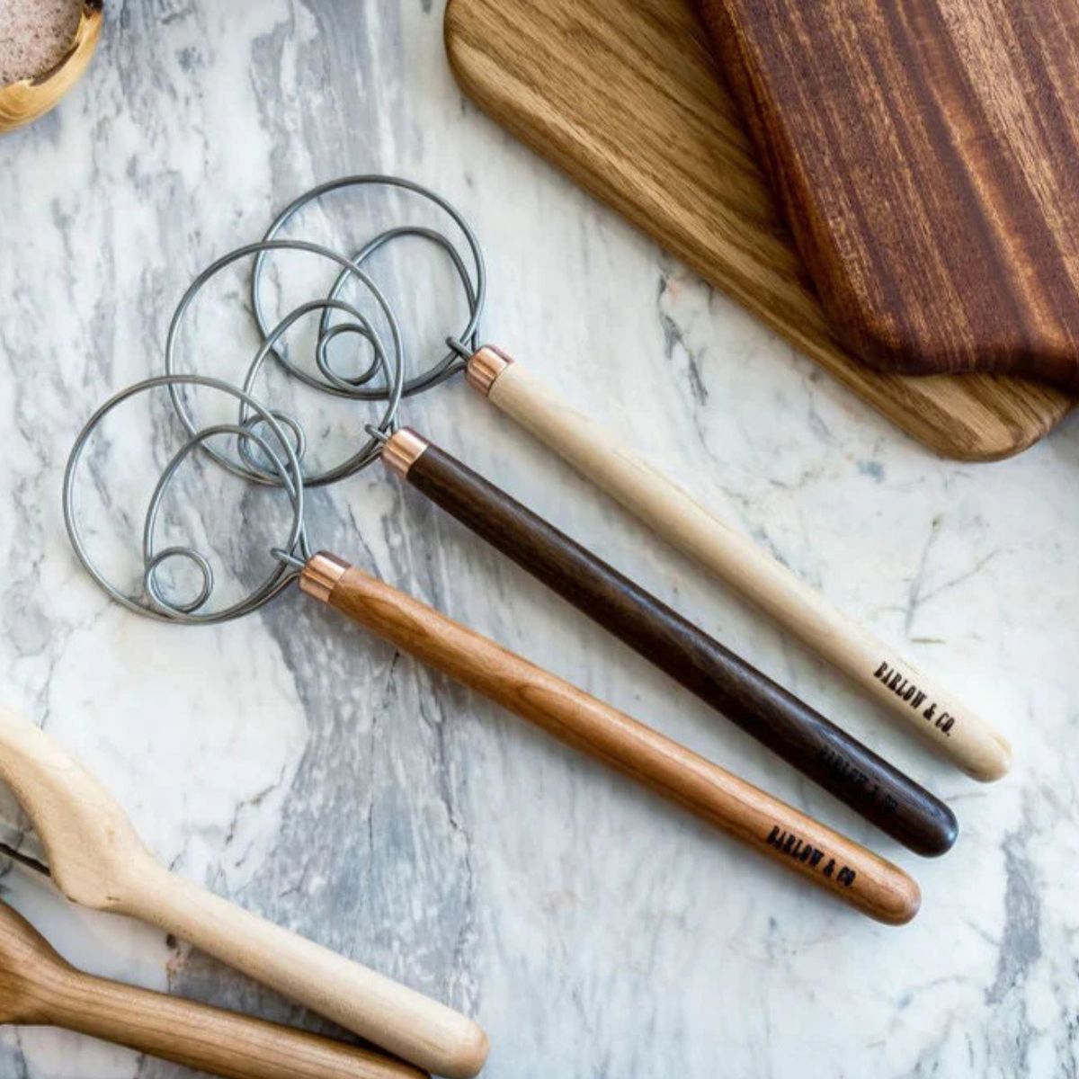 3 dough whisks in 3 different wood colors on a backdrop. 