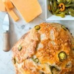 A loaf of Jalapeno Cheddar Sourdough Bread next to a bowl of pickled jalapenos and a block of cheddar cheese