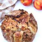 A freshly baked loaf of Peach Crisp Sourdough bread next to a white and brown napkin and fresh peaches.