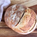 A loaf of Sourdough Bread next to a white and brown napkin on a wooden board.