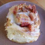 Single Sourdough Maple Cinnamon Roll on a purple plate with crumbled bacon on top.