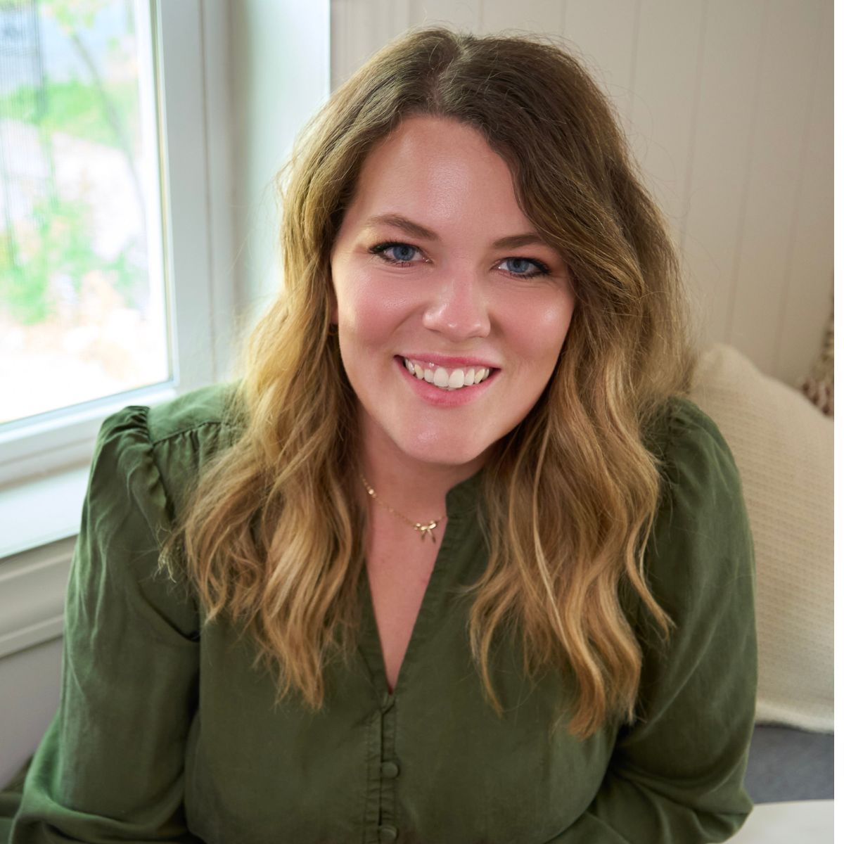 Emily Christensen from Country Roads Sourdough in a green dress, blonde hair and blue eyes with a window behind her.