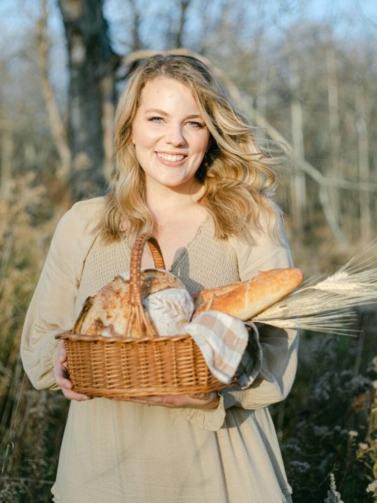 girl in a beige dress standing in a field with trees behind with a basket of bread