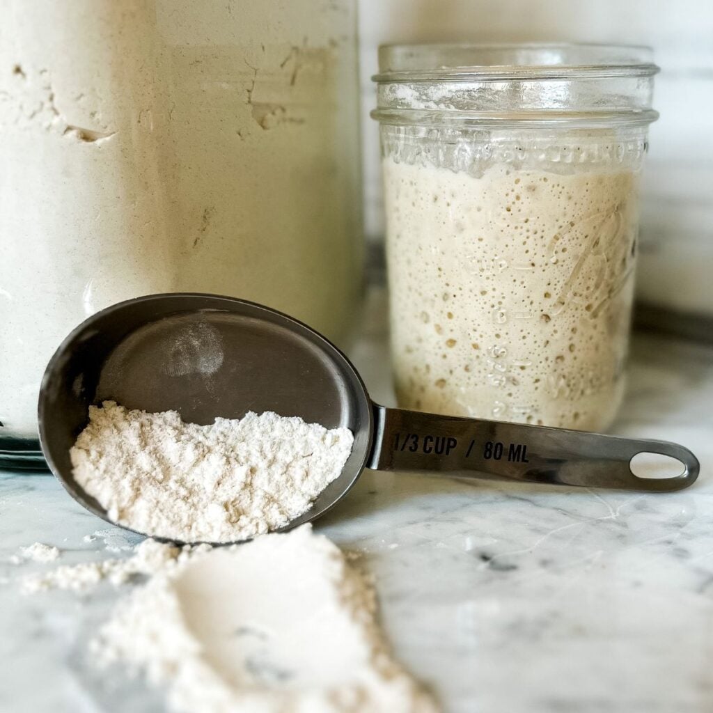 jar of sourdough starter next to a container of flour and a cup of flour with half of it dumped on the counter