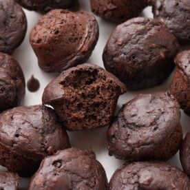A sourdough chocolate mini muffin with a bite taken out of it next to more mini muffins on a white surface.