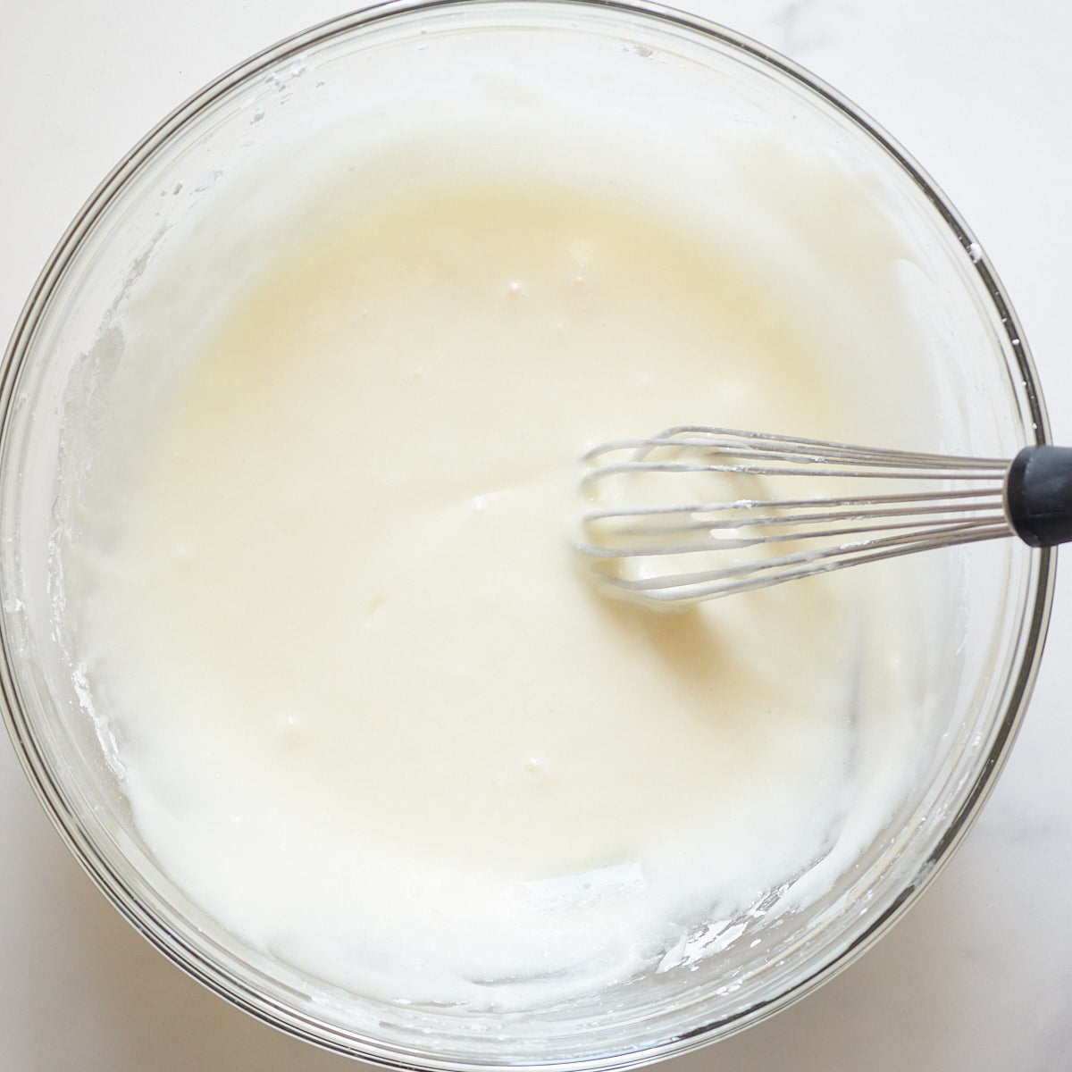 Clear bowl with cream cheese frosting and a small whisk.
