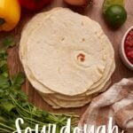 Pile of sourdough tortillas with peppers, onion, lime, salsa, and cilantro around it.