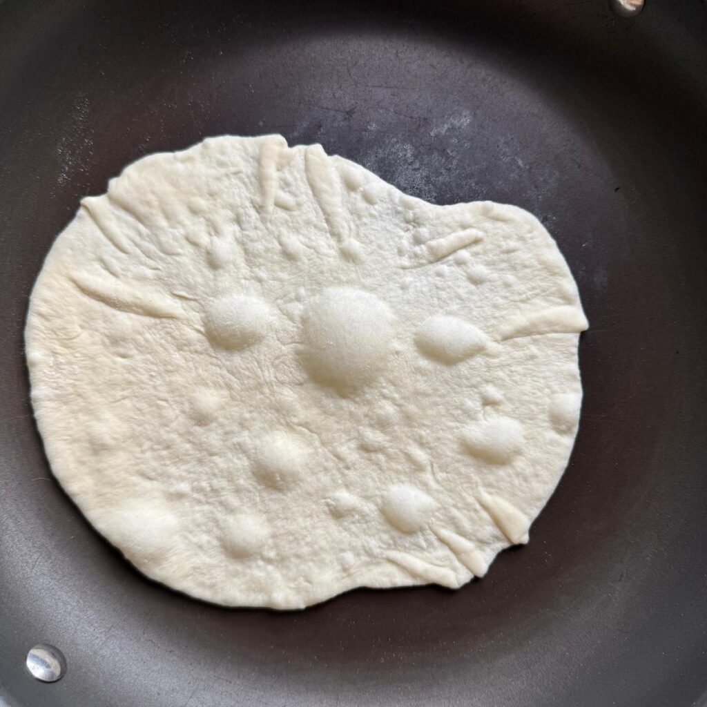 A homemade sourdough flour tortilla in a non-stick pan bubbling up before flipping it over. 