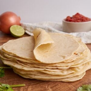Stack of flour tortillas with the top one rolled halfway with a lime, onion, and salsa in the background.
