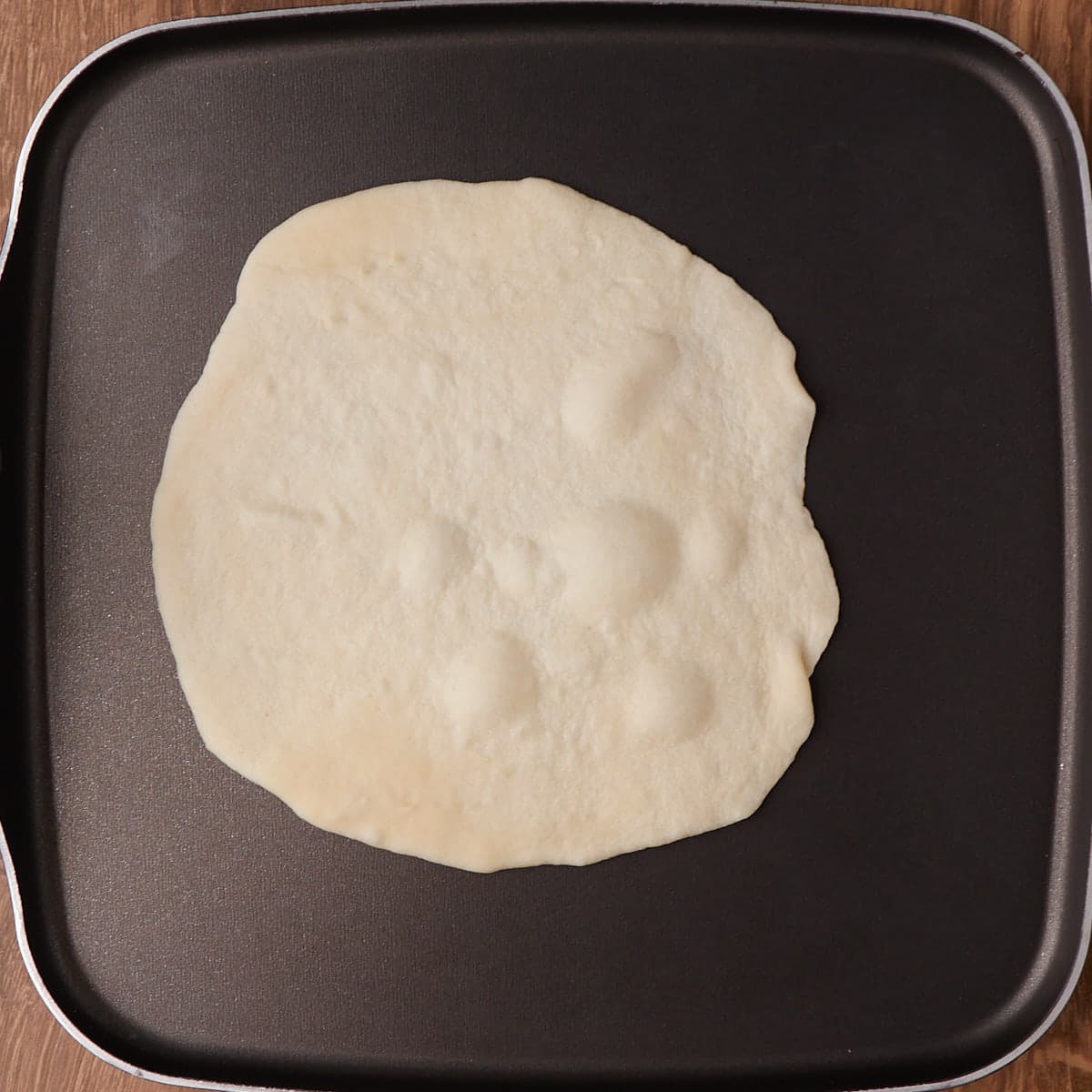 Sourdough tortilla on a black pan with bubbles coming up on the surface of the tortilla. 