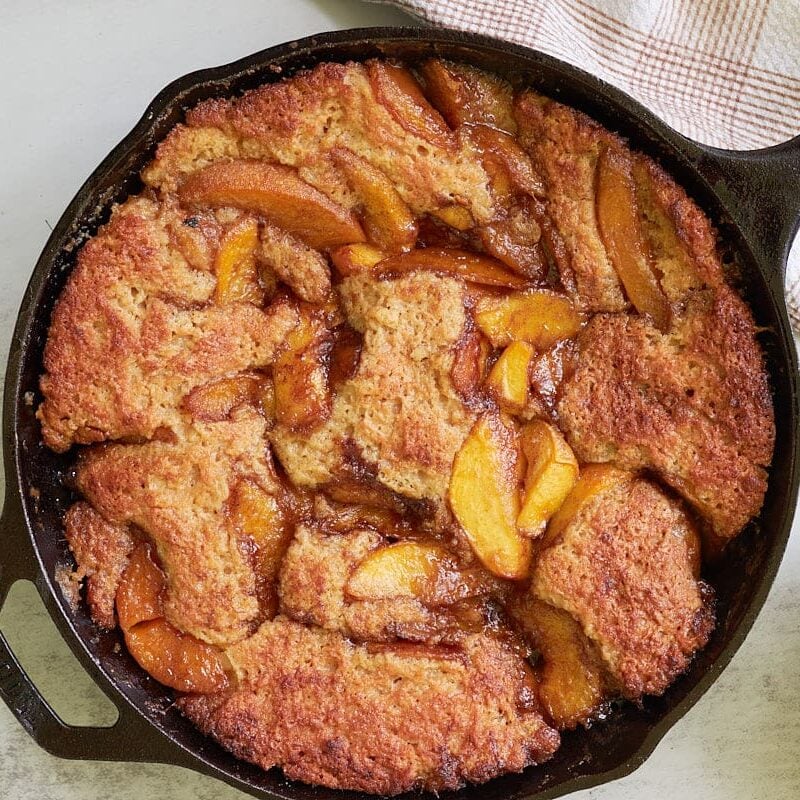 Freshly baked sourdough peach cobbler next to a bowl of peaches and a white and brown towel.