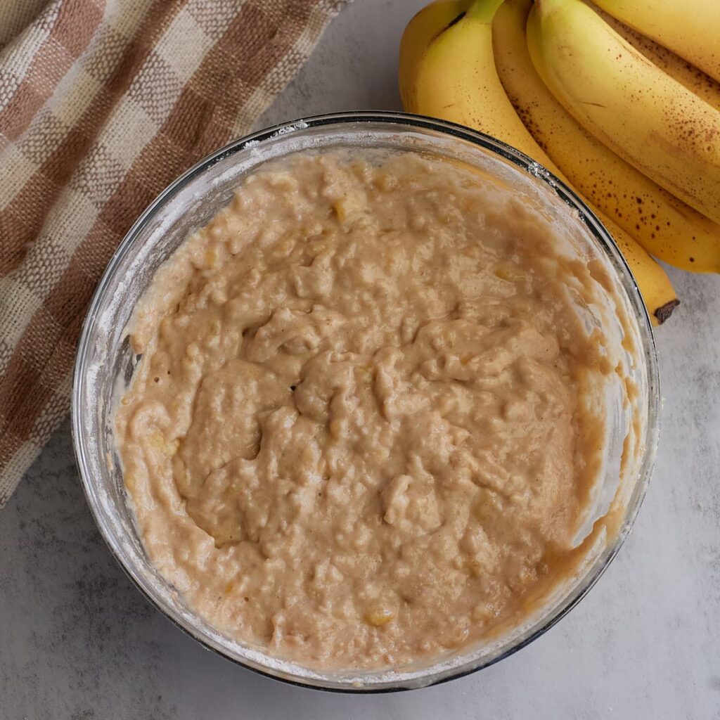 Batter for Sourdough Banana Muffins in a clear bowl next to a bunch of bananas.