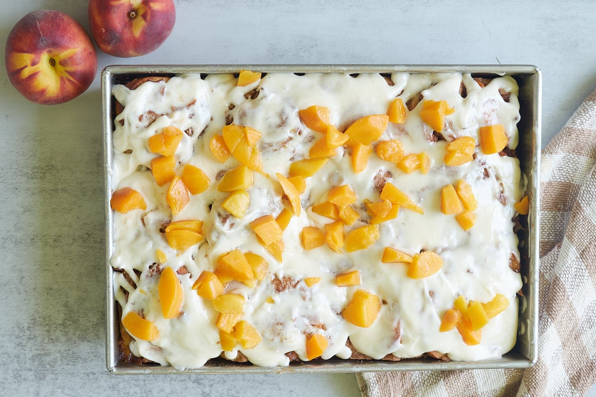 Sourdough peach cobbler cinnamon rolls in a 9x13 pan next to fresh peaches and a brown and white towel.