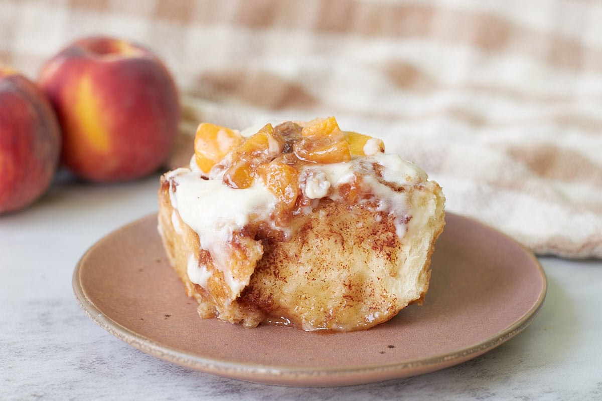 Sourdough peach cobbler cinnamon roll on a brown plate next to fresh peaches.