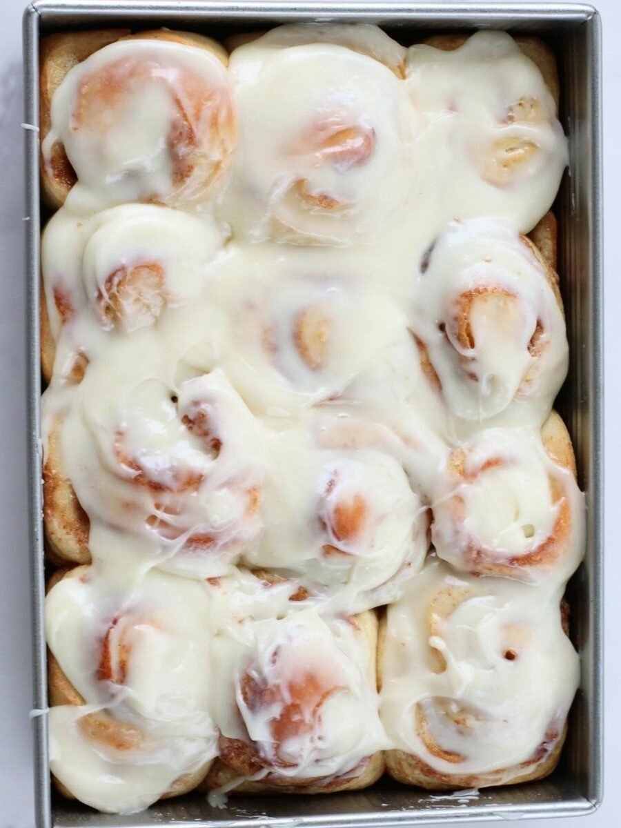 cream cheese icing covering 12 cinnamon rolls in a pan