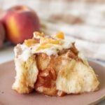 Sourdough peach cobbler cinnamon roll on a brown plate next to fresh peaches.