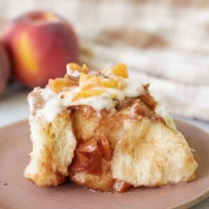Sourdough peach cobbler cinnamon roll on a brown plate next to fresh peaches.