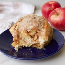 Apple Crisp Cinnamon Roll on a blue plate next to red apples.