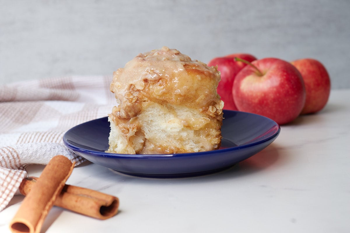 One Apple Crisp Sourdough Cinnamon Roll on a blue plate next to red apples and cinnamon sticks.