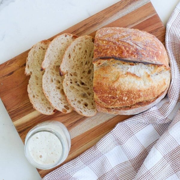 Same Day Sourdough Bread sliced on a wooden cutting board next to a brown and white cloth napkin and a jar of active sourdough starter.