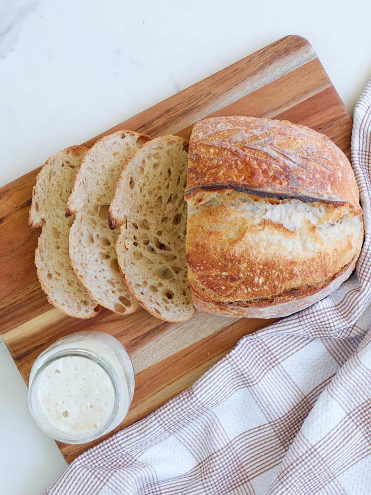 Same Day Sourdough Bread sliced on a wooden cutting board next to a brown and white cloth napkin and a jar of active sourdough starter.