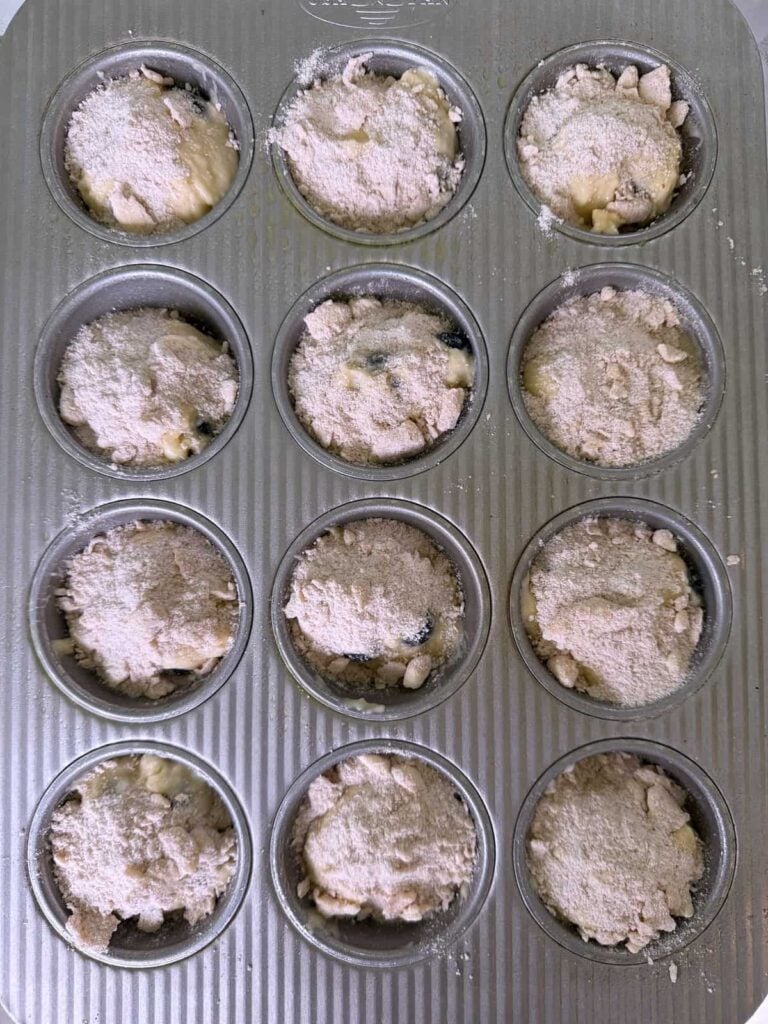 Streusel added on top of unbaked sourdough blueberry muffin matter in a metal muffin tin.