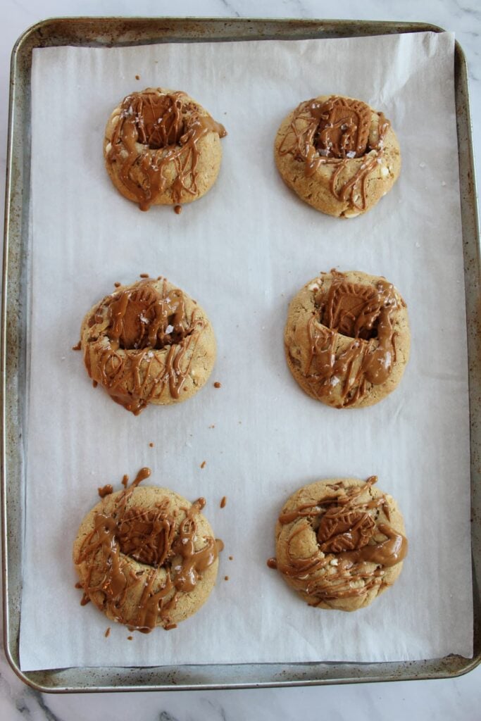 Freshly baked cookie butter sourdough cookies with a biscoff cookie inserted into the center and a cookie butter drizzle on a parchment lined sheet pan.