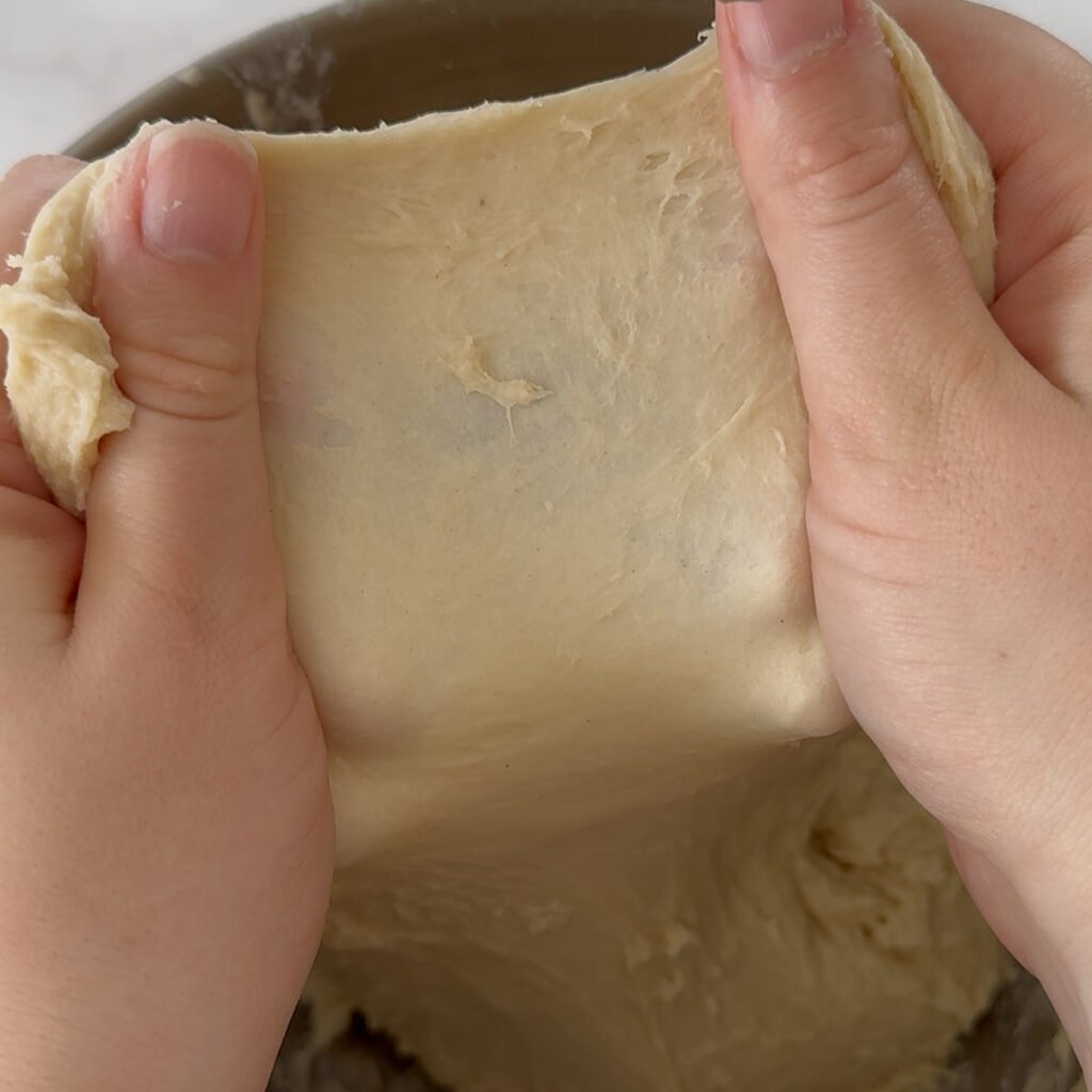 Hand holding out a piece of dough doing the windowpane test for sourdough dinner rolls.