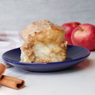 an apple crisp sourdough cinnamon roll on a blue plate with cinnamon sticks in front and apples behind.