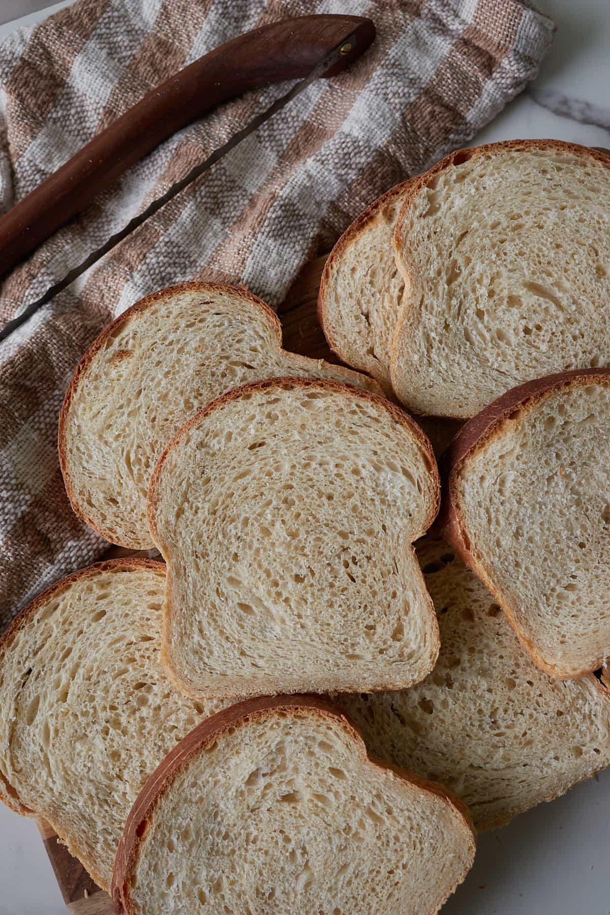 Slices of Sourdough Sandwich Bread on top of each other next to a brown and white towel and bread slicer.