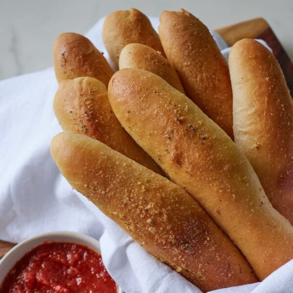 Sourdough breadsticks in a white lined bowl with a white cup of marinara sauce.