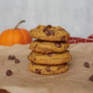 A stack of 4 sourdough pumpkin chocolate chip cookies on a piece of parchment paper with a pumpkin in the background.