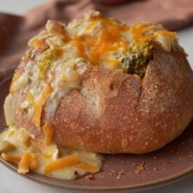 A sourdough bread bowl with broccoli and cheddar soup with cheese on top on a brown plate.
