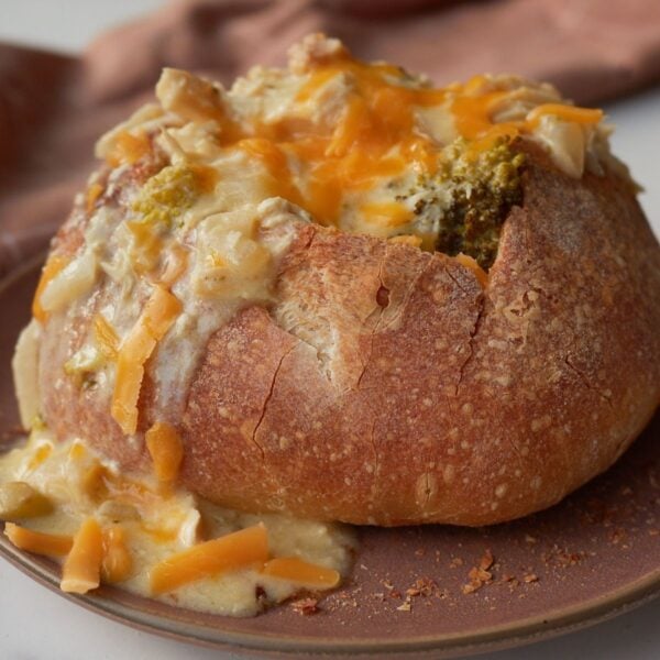A sourdough bread bowl with broccoli and cheddar soup with cheese on top on a brown plate.