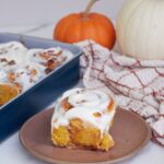 Sourdough pumpkin cinnamon roll on a brown plate next to a 9x13 pan with rolls and an orange and white pumpkin.