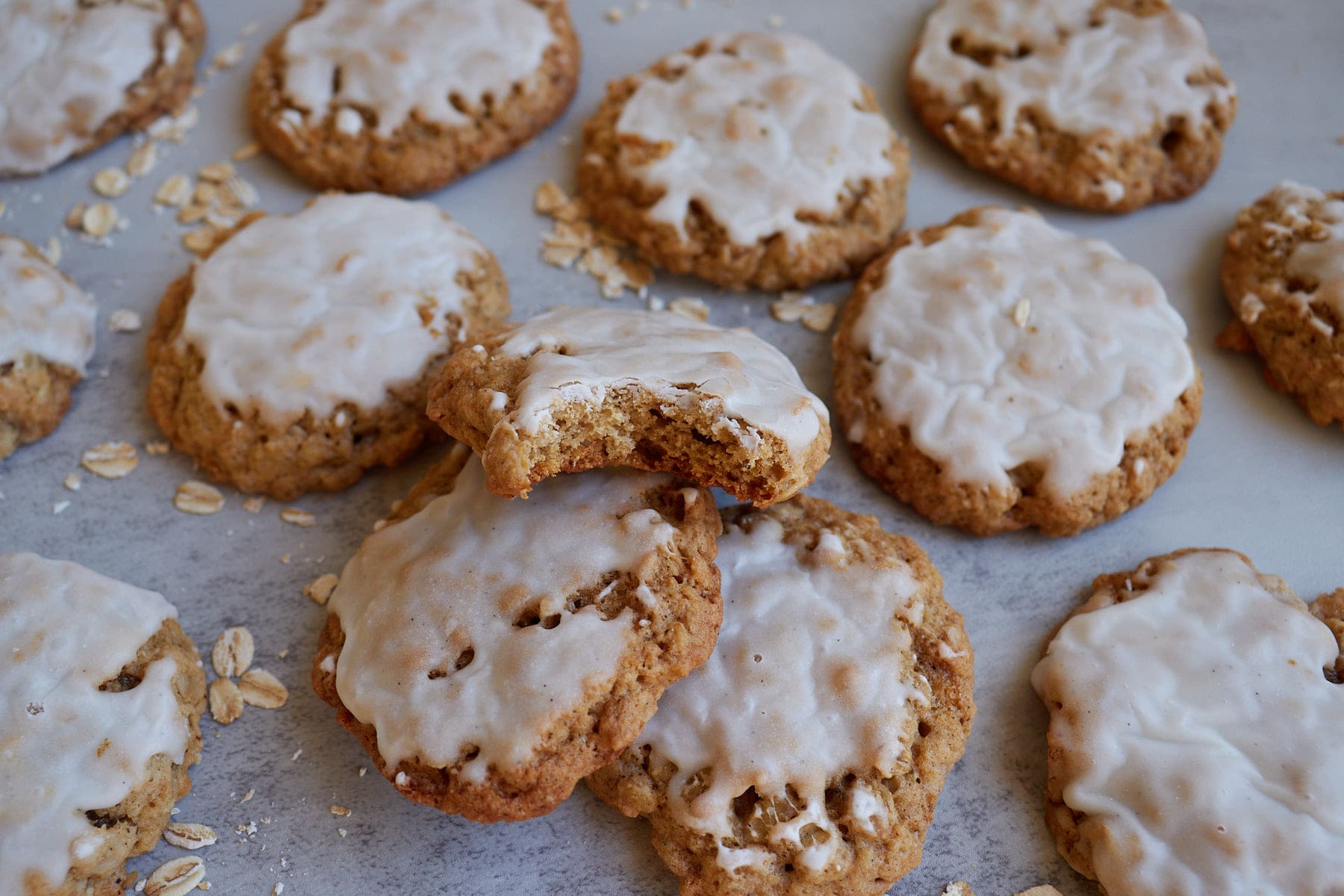 Sourdough Oatmeal Cookies with a vanilla bean glaze on a grey background with oats scattered and one cookie with a bite taken out of it.