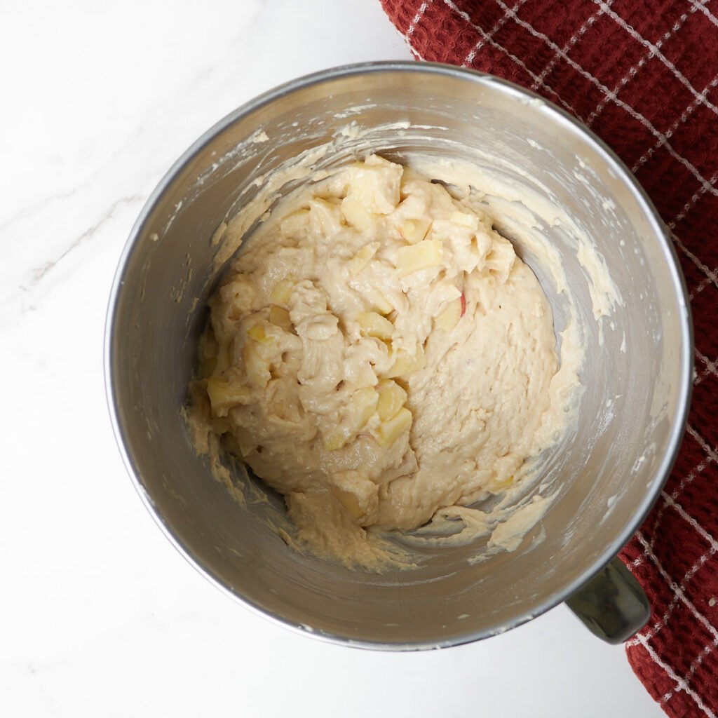 Apples folded into batter for Sourdough Apple Fritter Bread in a bowl of stand mixer next to a red towel.