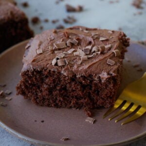 Slice of chocolate cake with chocolate butter cream frosting and chocolate shavings on a brown plate.