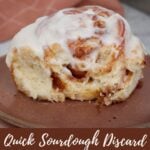 One sourdough discard cinnamon roll on a brown plate with a bite taken out of it next to a 9x13 pan filled with sourdough discard cinnamon rolls.