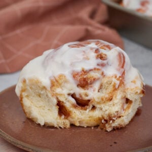 One sourdough discard cinnamon roll on a brown plate with a bite taken out of it next to a 9x13 pan filled with sourdough discard cinnamon rolls.