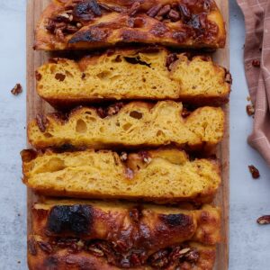 3 slices of sourdough sweet potato focaccia on their sides on a wooden cutting board with pecans scattered around.