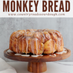 A Pinterest pin showcasing sourdough monkey bread with icing dripping off the top featured on a beautiful wooden cake stand. The cake stand is on a white marble counter with a dark marble background. A white cloth is on the counter next to the cake stand.