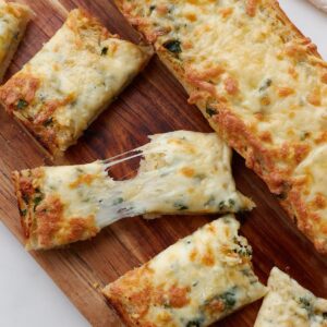 Slices of cheesy garlic bread on a wood cutting board with one of the slices pulled apart with cheesy strands holding it together.