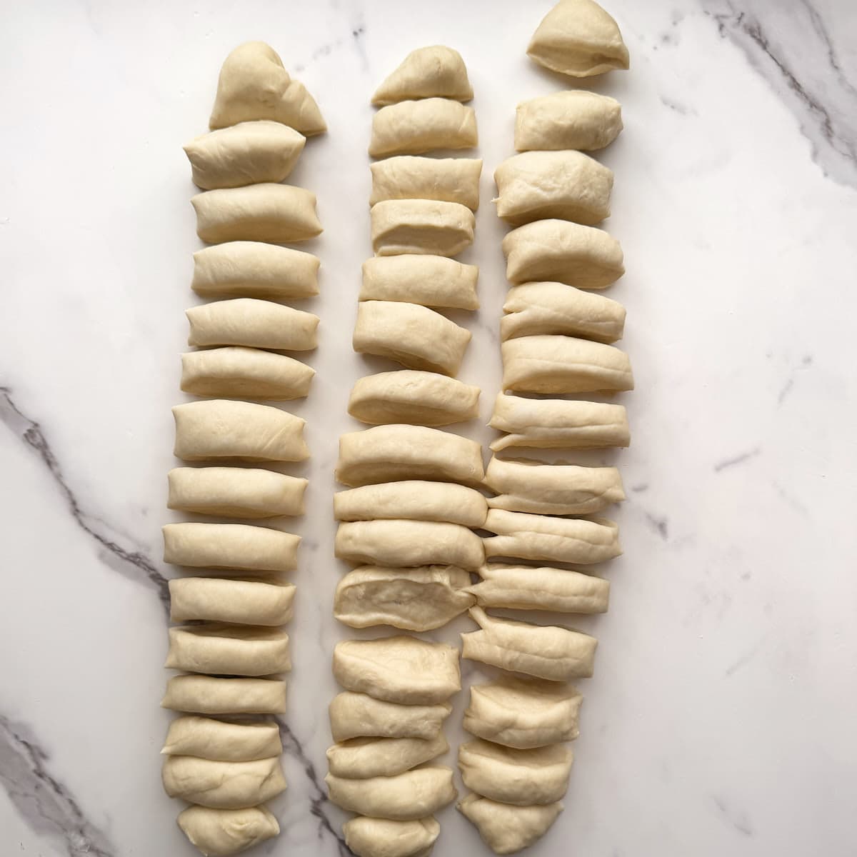 Sourdough monkey bread dough divided into three equal parts and rolled into 15" logs and cut into 1" pieces on a marble counter.